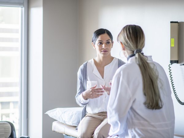 A young female of Indian ethnicity sits on the examination table at her gynaecologist's office. She is talking to her doctor about her health history.
