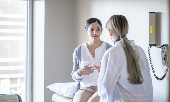 A young female of Indian ethnicity sits on the examination table at her gynaecologist's office. She is talking to her doctor about her health history.