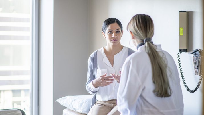 A young female of Indian ethnicity sits on the examination table at her gynaecologist's office. She is talking to her doctor about her health history.