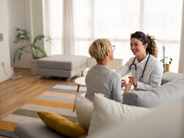 Caucasian female physician conducts friendly medical consultation with senior patient, wearing white coat and stethoscope during residential healthcare visit.