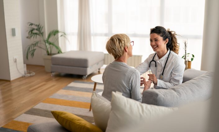 Caucasian female physician conducts friendly medical consultation with senior patient, wearing white coat and stethoscope during residential healthcare visit.