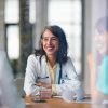 A friendly female doctor wearing a stethoscope sits at a table, engaging with patients in a glass-walled hospital setting. Bright light and professional teamwork convey trust, care, and medical collaboration.